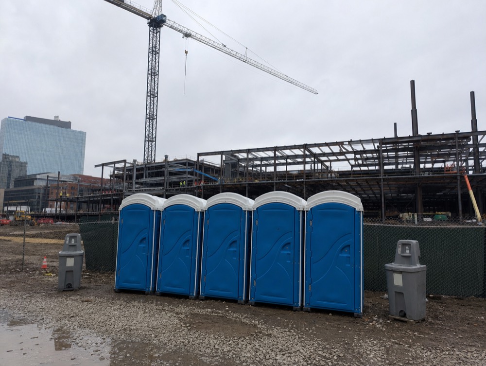 Five portable toilets and two hand wash stations at a steel-frame construction site on an overcast day
